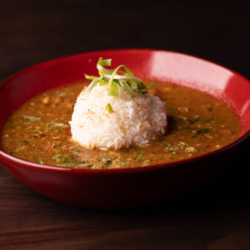 A red bowl of Southern gumbo with a mound of white rice and green onions on a dark wooden table.