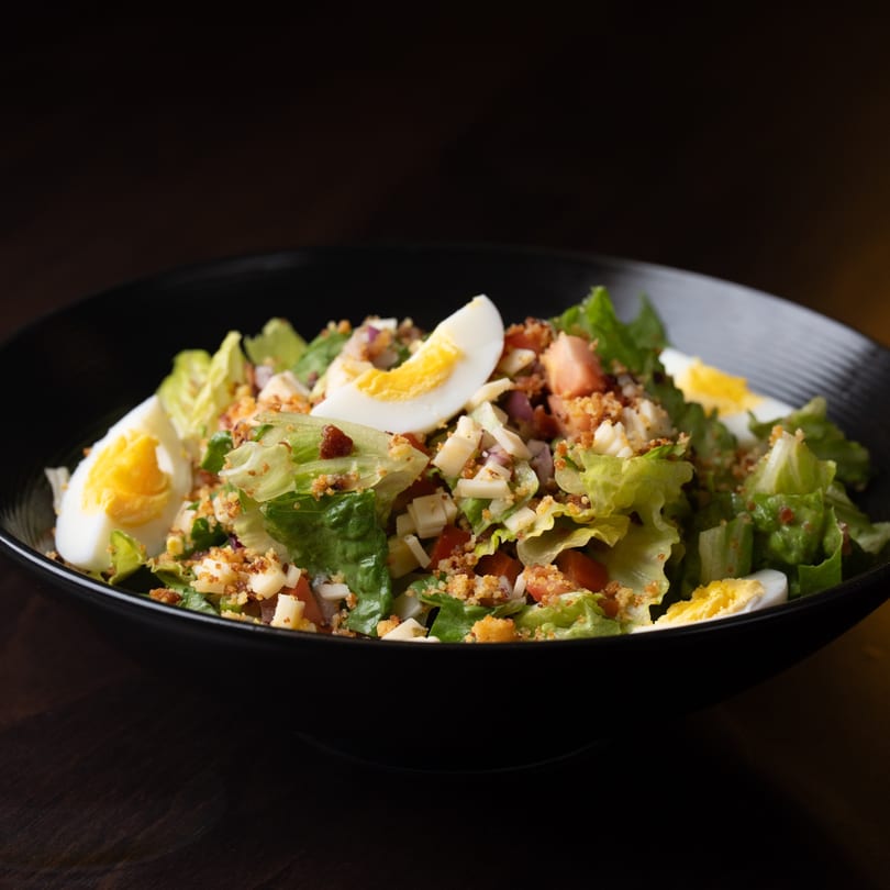A Southern-style salad with lettuce, hard-boiled eggs, cheese, tomatoes, and bacon bits in a black bowl.