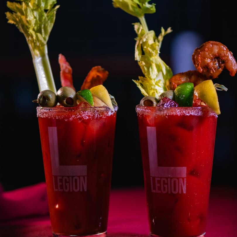 Two Legion Sports Bar Bloody Marys, garnished with celery, olives, lime, lemon, and shrimp.