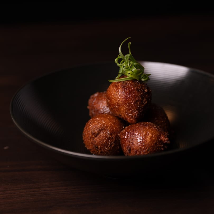 A black bowl filled with golden-brown hush puppies, garnished with green scallions, on a dark wooden surface.